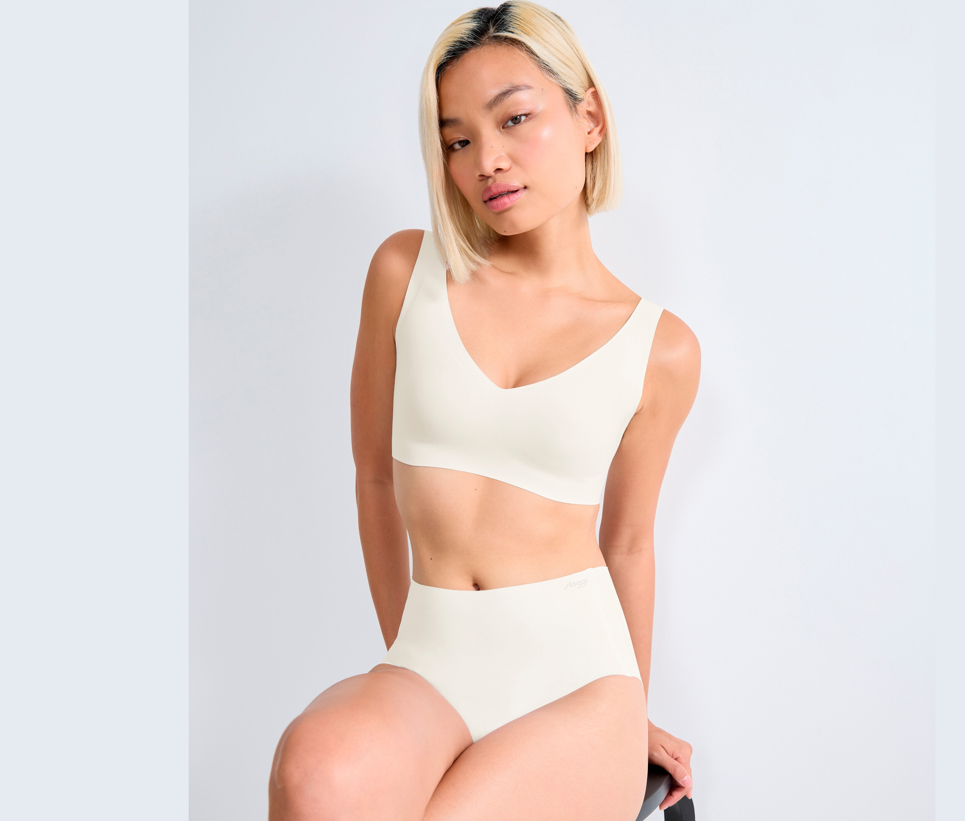 Kobieta siedzi i ma na sobie biały biustonosz bralette sloggi ZERO Feel 2.0 i majtki.