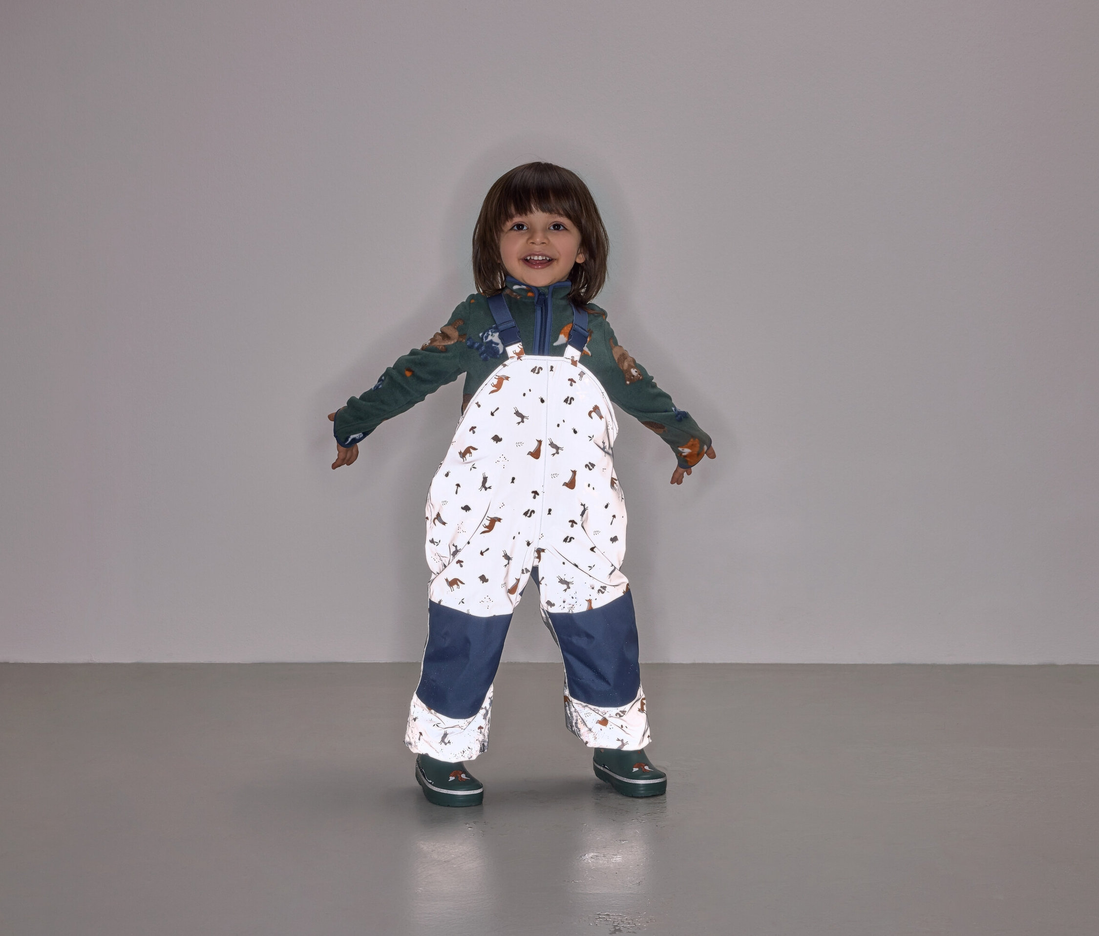 Uśmiechnięte dziecko stoi z rozłożonymi ramionami, ubrane w kombinezon na szelkach z nadrukiem zwierząt i granatowymi nogawkami.