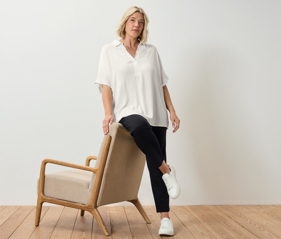 Blondwłosa kobieta pozuje oparta o beżowy fotel, ma na sobie kremową bluzkę.