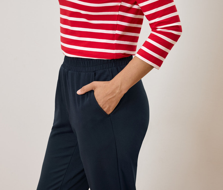 Kobieta z ręką w kieszeni granatowych spodni i czerwono-białej bluzce w paski.