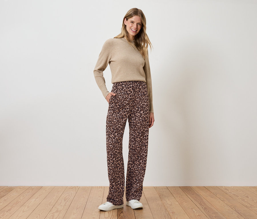 Kobieta stoi z rękami w kieszeniach brązowych kwiecistych spodni i beżowym swetrze.