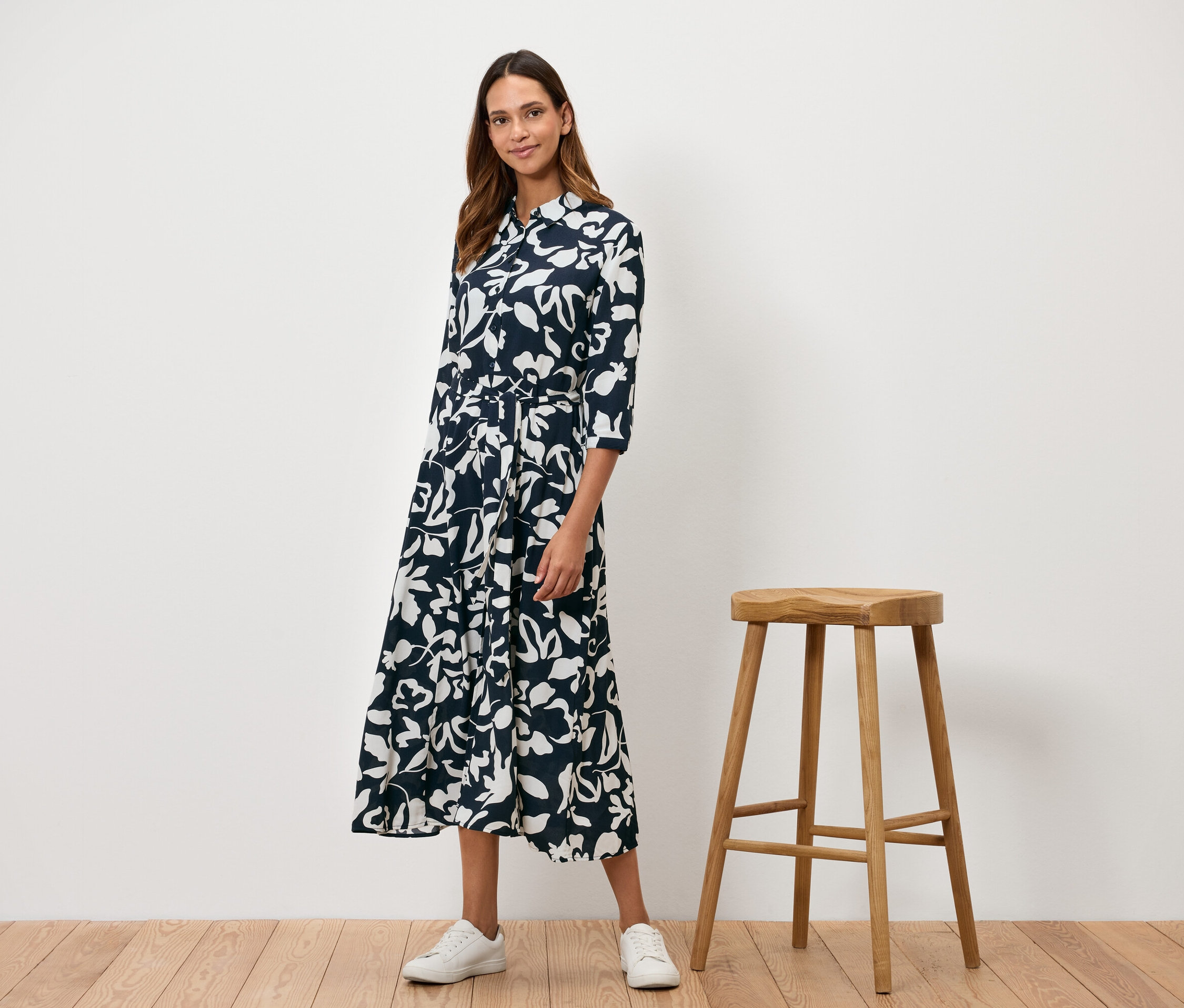 Kobieta stoi obok drewnianego stołka i ma na sobie granatową sukienkę w białe kwiaty.