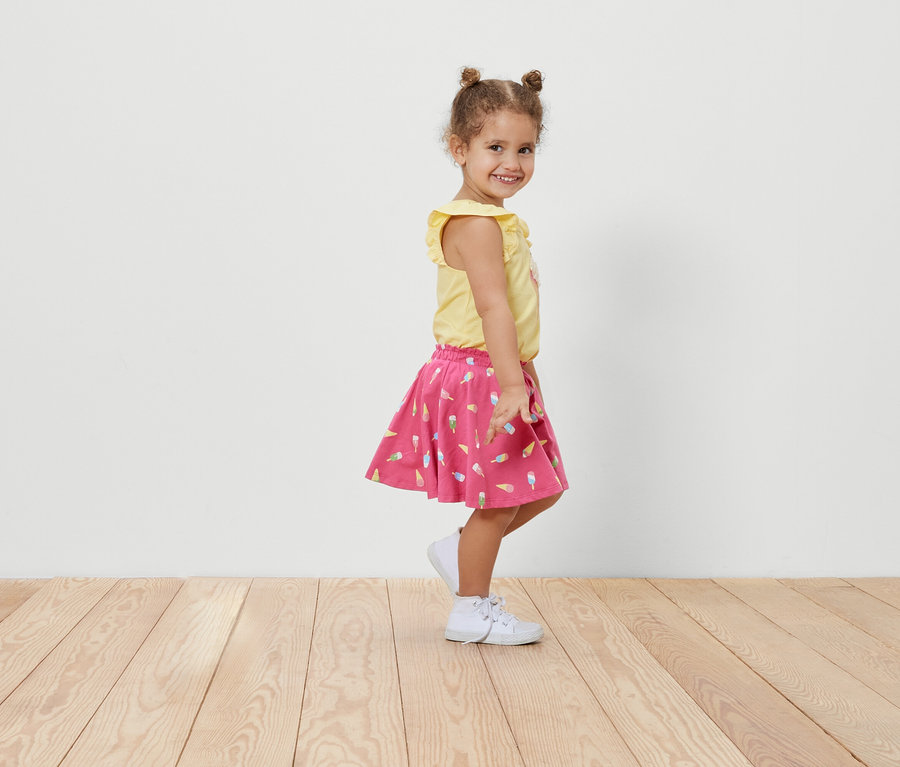 Uśmiechnięta dziewczynka pozuje bokiem w spódnicy z dżerseju ze spodenkami we wzór lodów i żółtej koszulce.