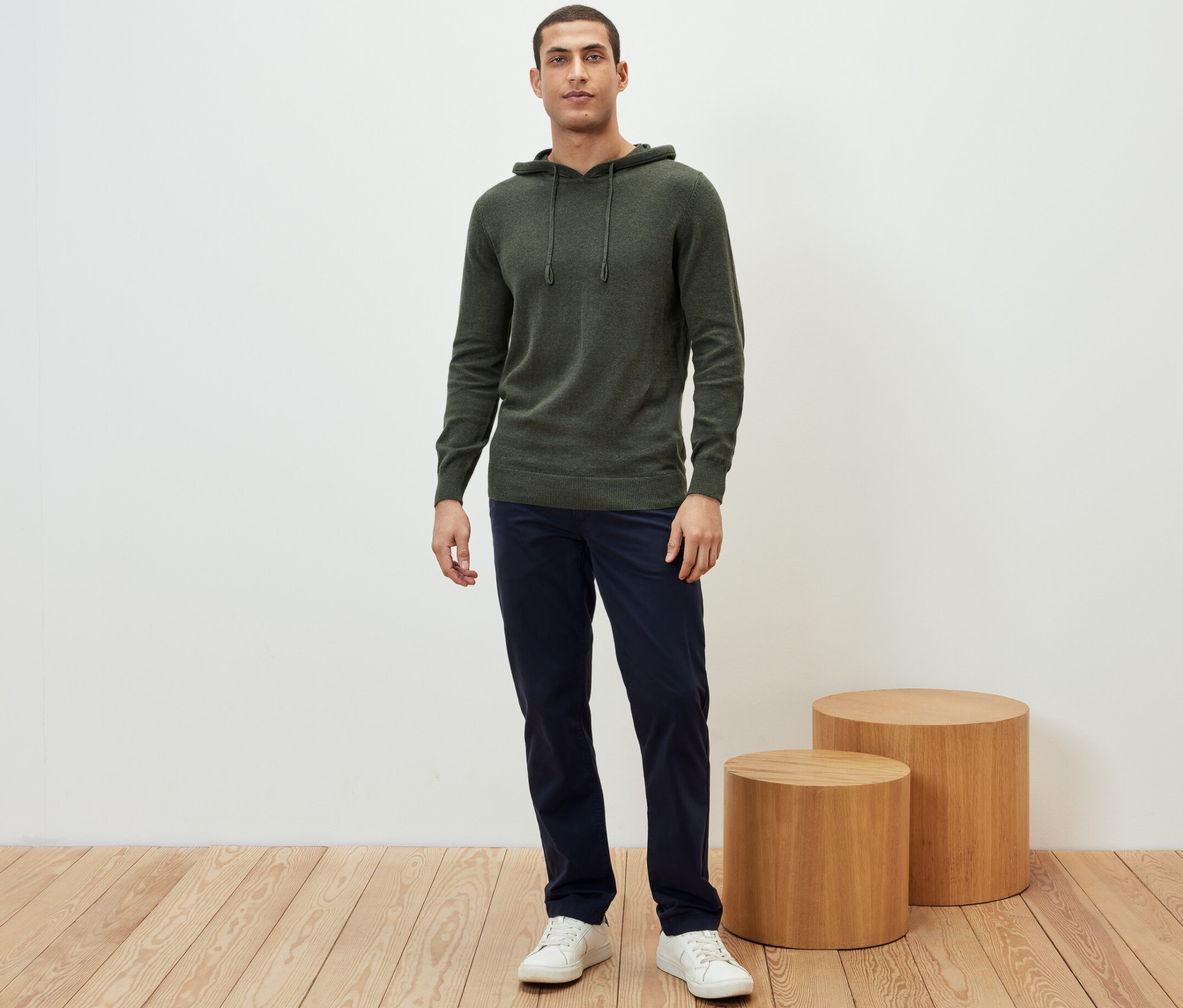 Mężczyzna stoi w dzianinowej bluzie z kapturem, granatowych spodniach i białych tenisówkach.