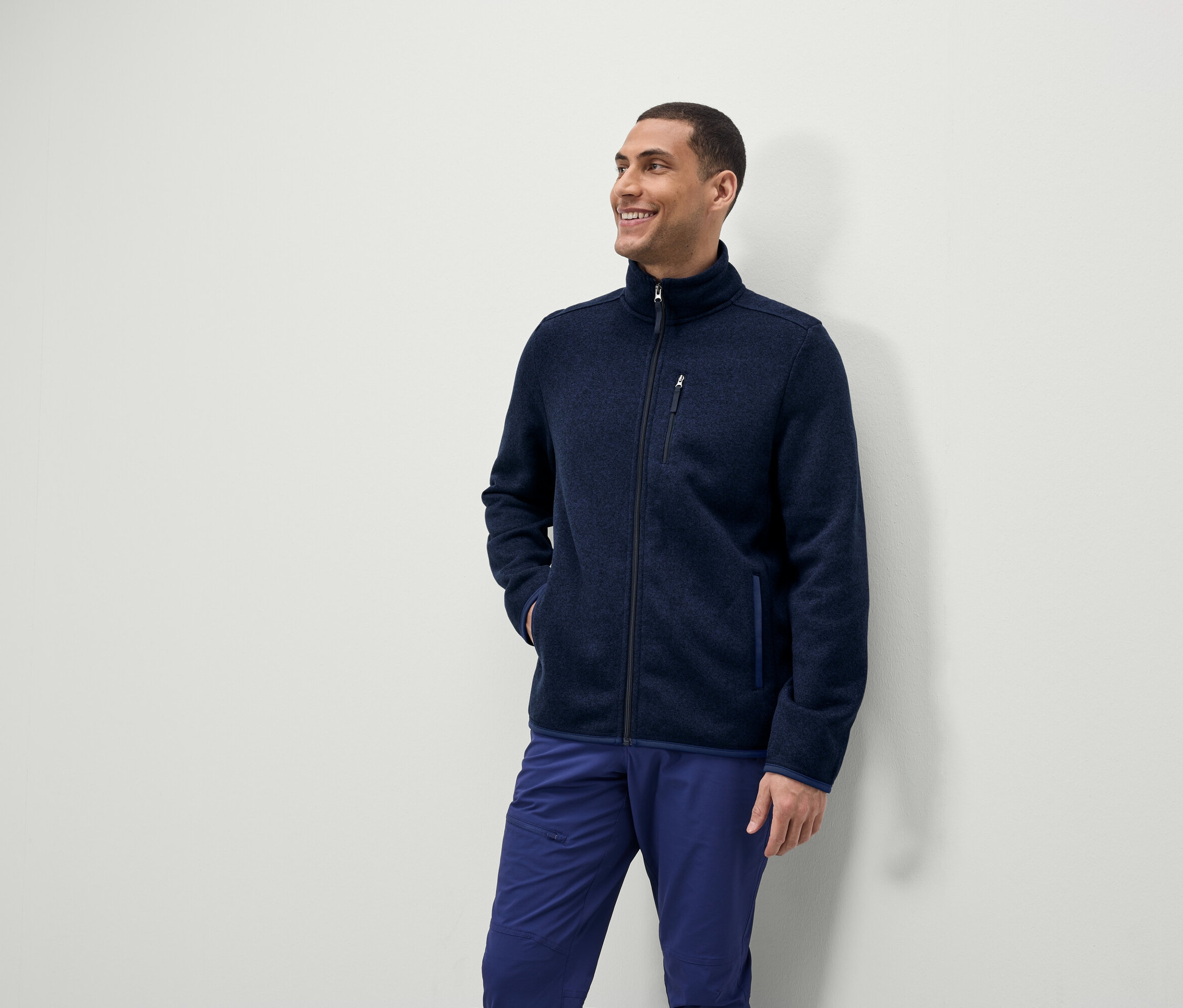 Mężczyzna uśmiecha się i patrzy w bok, ubrany w niebieską bluzę polarową na zamek i niebieskie spodnie.