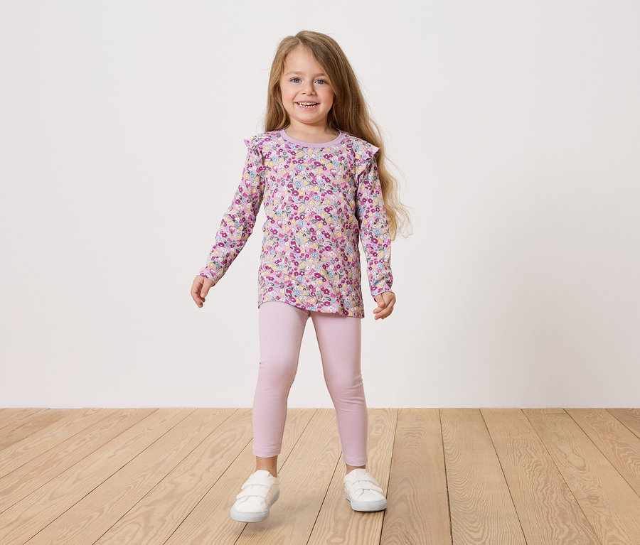 Dziewczynka z blond włosami ma na sobie bluzkę w kwiaty, różowe legginsy i białe buty.