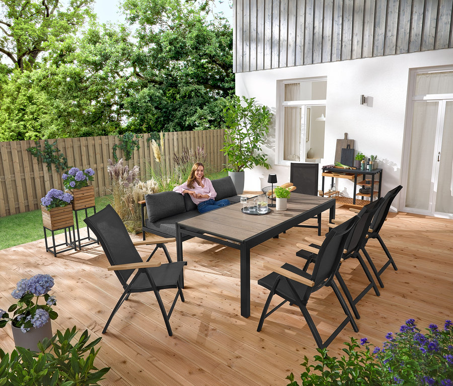 Kobieta siedzi na ławce ogrodowej 2 w 1 ‒ leżance »Elin«. W tle widoczne kolumny na rośliny „Tinus”, 2 sztuki. Na tarasie stoi również wózek do grillowania z powierzchnią roboczą z polikrylu i rozkładany stół ogrodowy z blatem Duraboard.