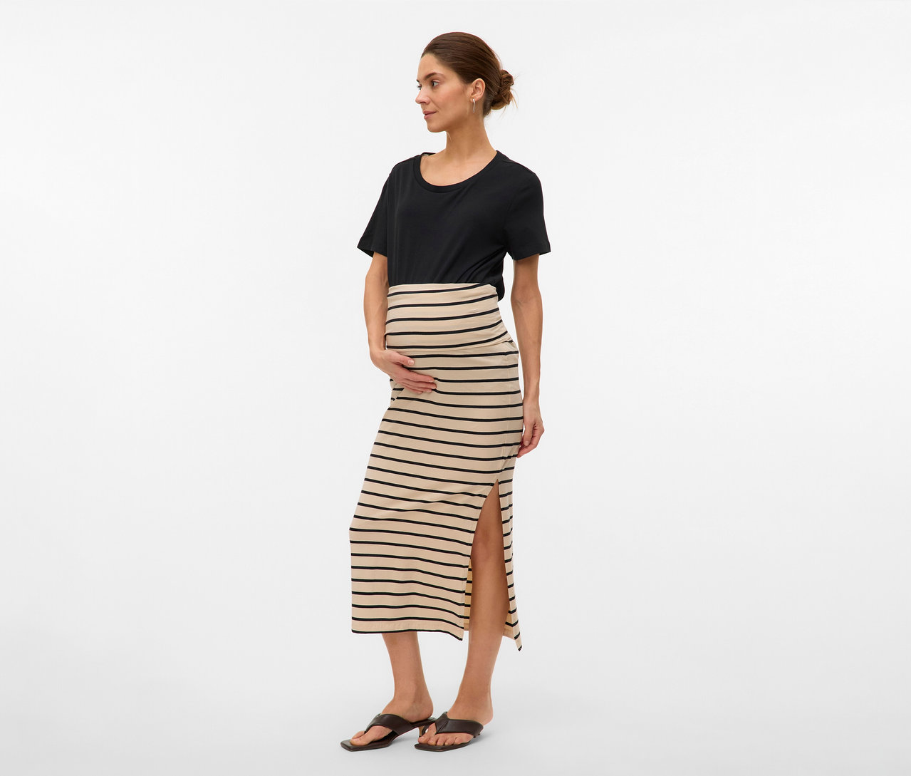 Kobieta w czarnym T-shircie i w pasiastą spódnicę maxi MAMALICIOUS „MLEVA” z rozcięciem. Trzyma ręce na brzuchu.