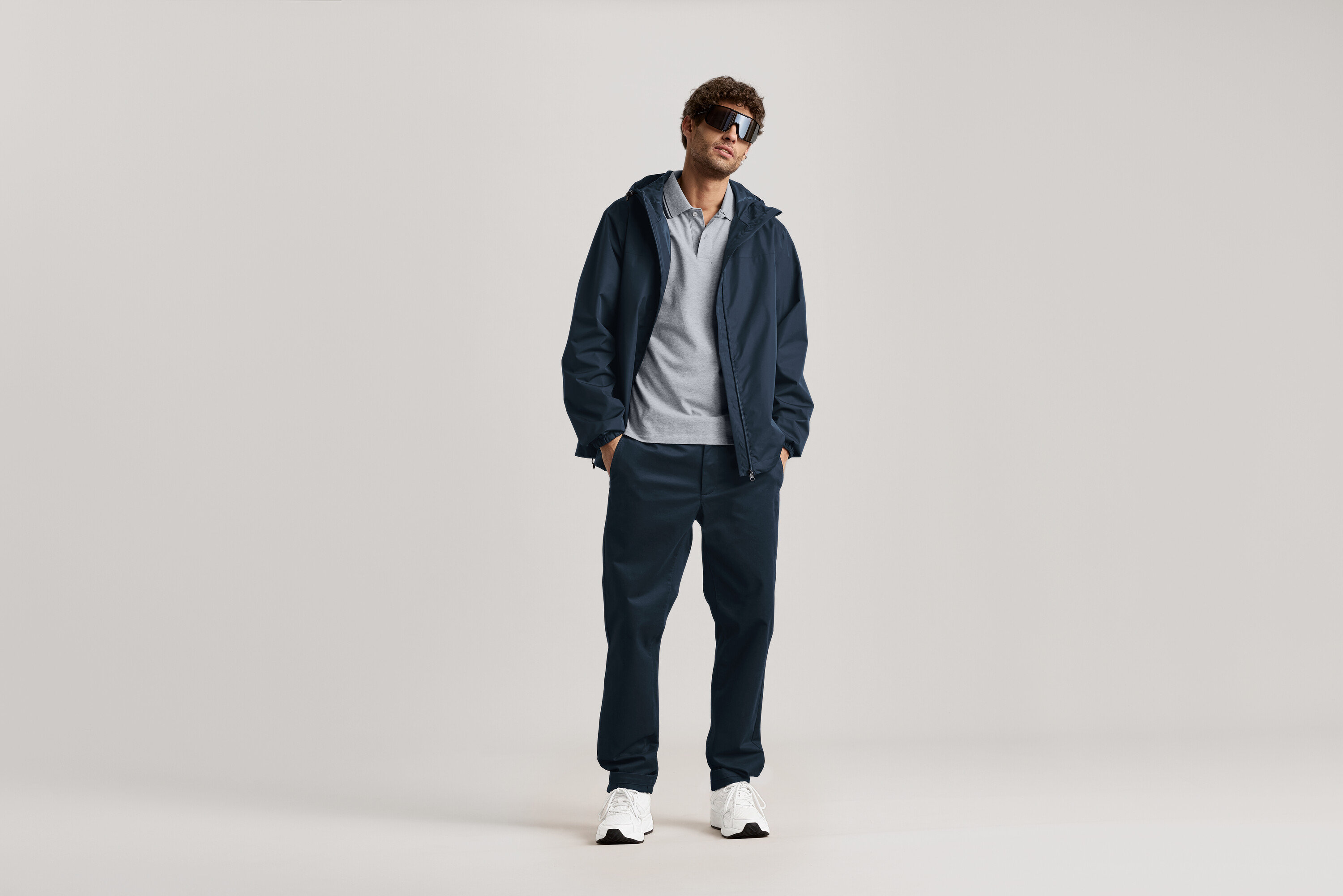 Mężczyzna stoi w okularach przeciwsłonecznych, granatowej kurtce i granatowych spodniach typu chino.