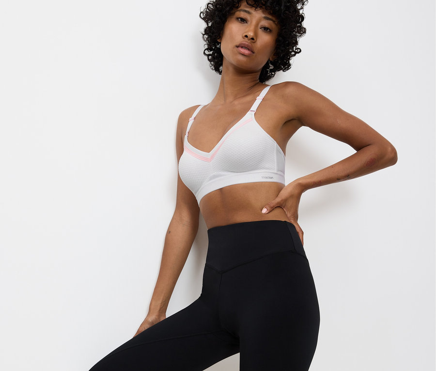 Kobieta pozuje w białym staniku sportowym z różowymi akcentami i czarnych legginsach na tle białej ściany.