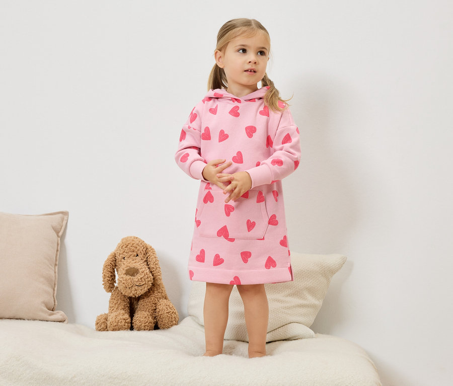 Dziewczynka z blond włosami w kucykach stoi na łóżku, ubrana w różową sukienkę z kapturem i czerwonymi sercami. Obok niej siedzi pluszowy pies.