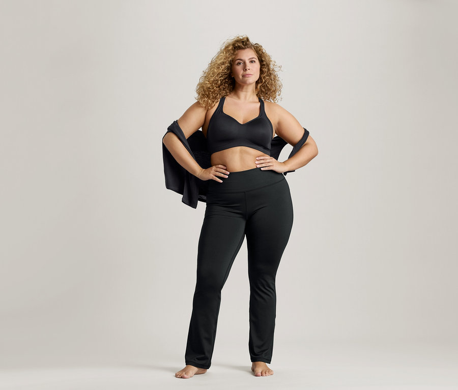 Kobieta o kręconych włosach w czarnym sportowym biustonoszu i legginsach, z kurtką na ramionach i rękami na biodrach.