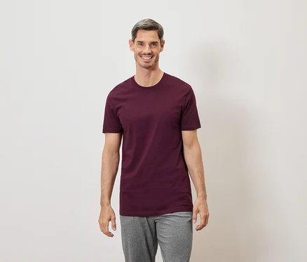 Uśmiechnięty mężczyzna z siwymi włosami w bordowym T-shircie i szarych spodniach na białym tle.