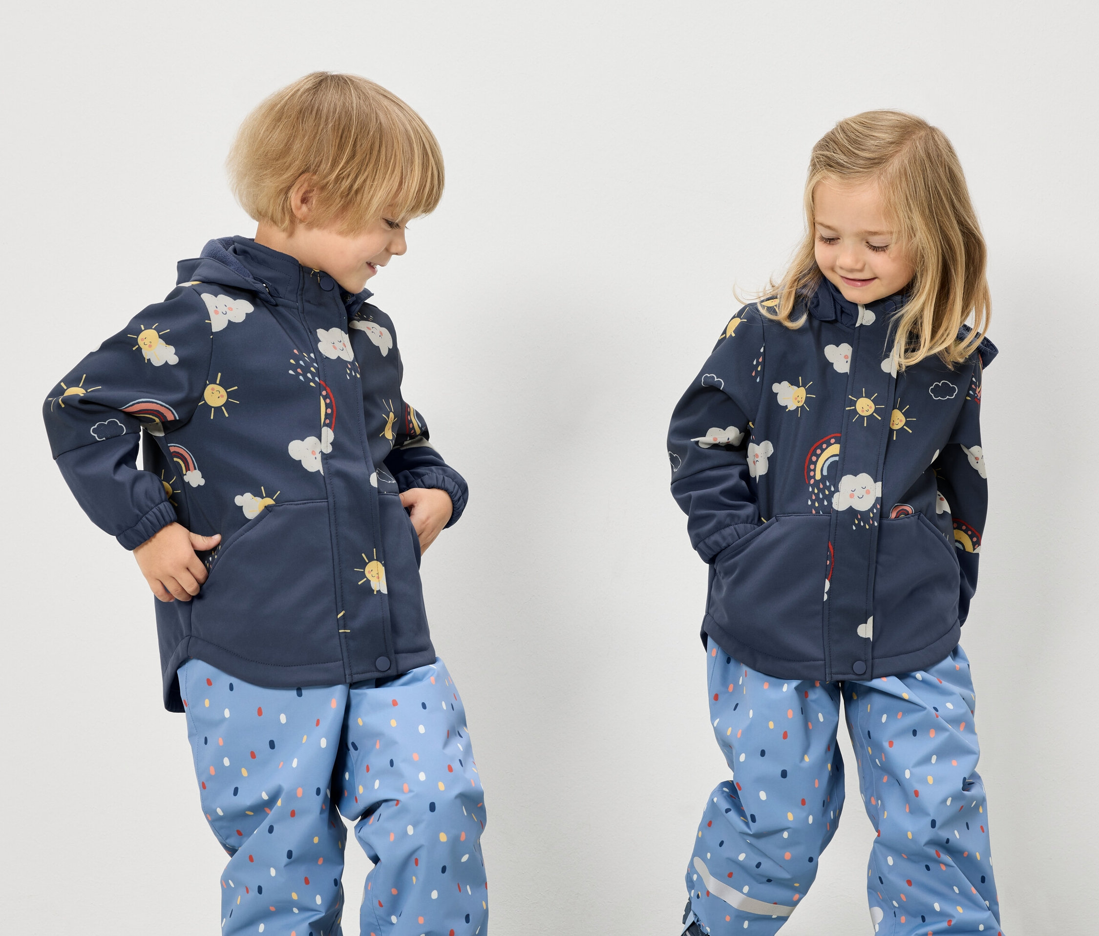 Dwoje dzieci w niebieskich kurtkach we wzory z tęczami, chmurami i słońcami oraz niebieskich spodniach w kropki stoi na białym tle.