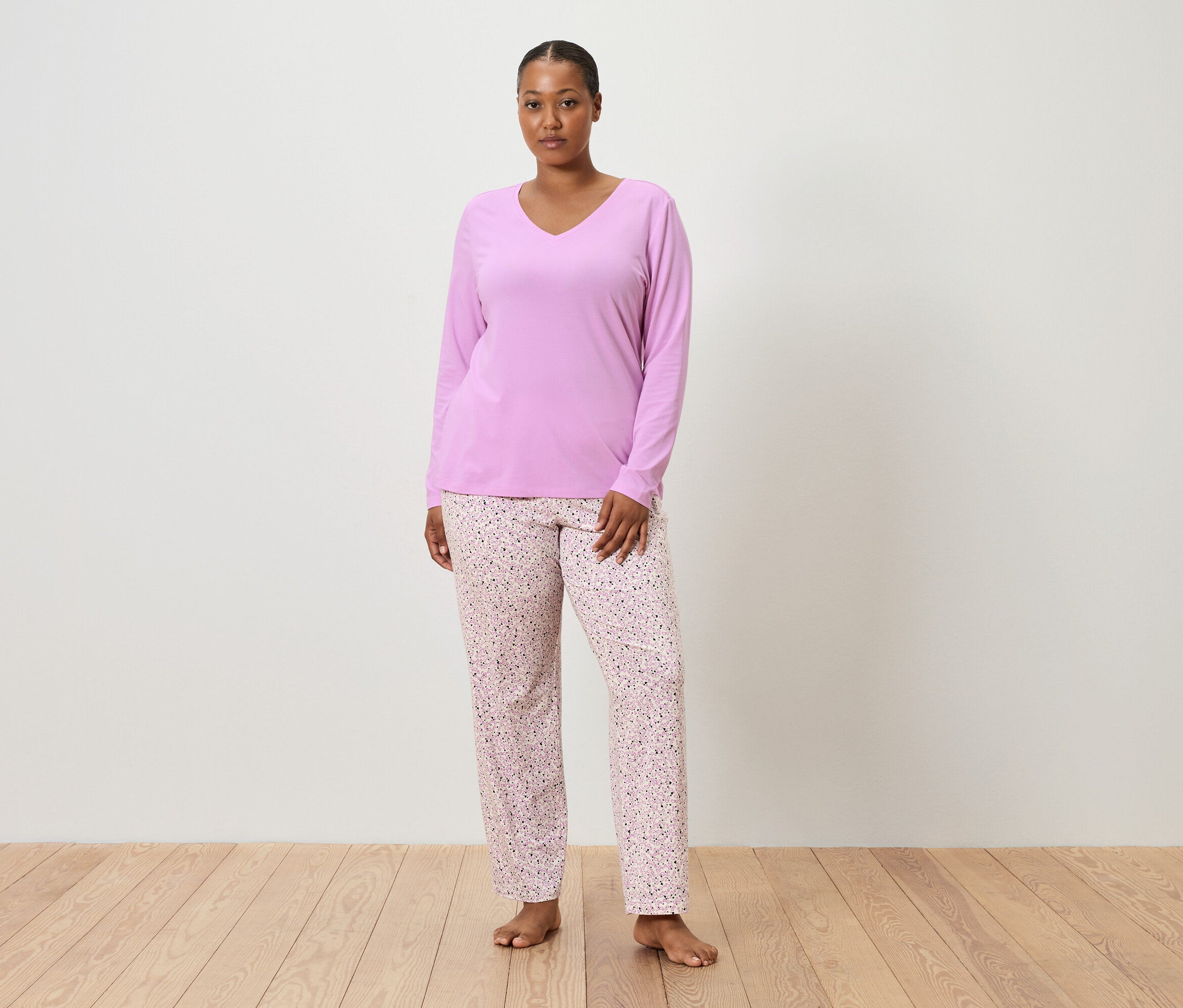 Kobieta stoi boso na drewnianej podłodze na tle białej ściany, ubrana w liliową bluzkę od piżamy i różowe spodnie od piżamy w kwiatowy wzór.