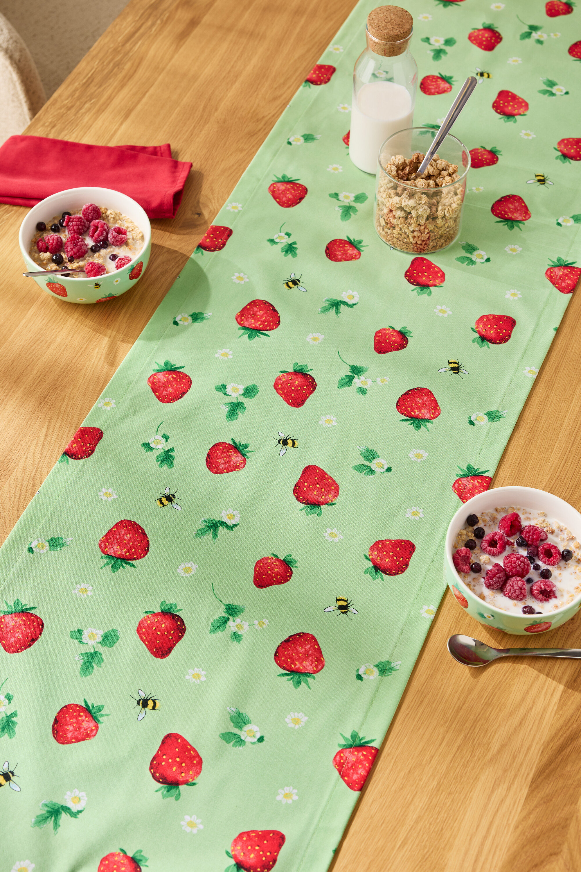 Na drewnianym stole leży bieżnik w truskawki, na nim miski z płatkami, szklanka musli i butelka mleka.