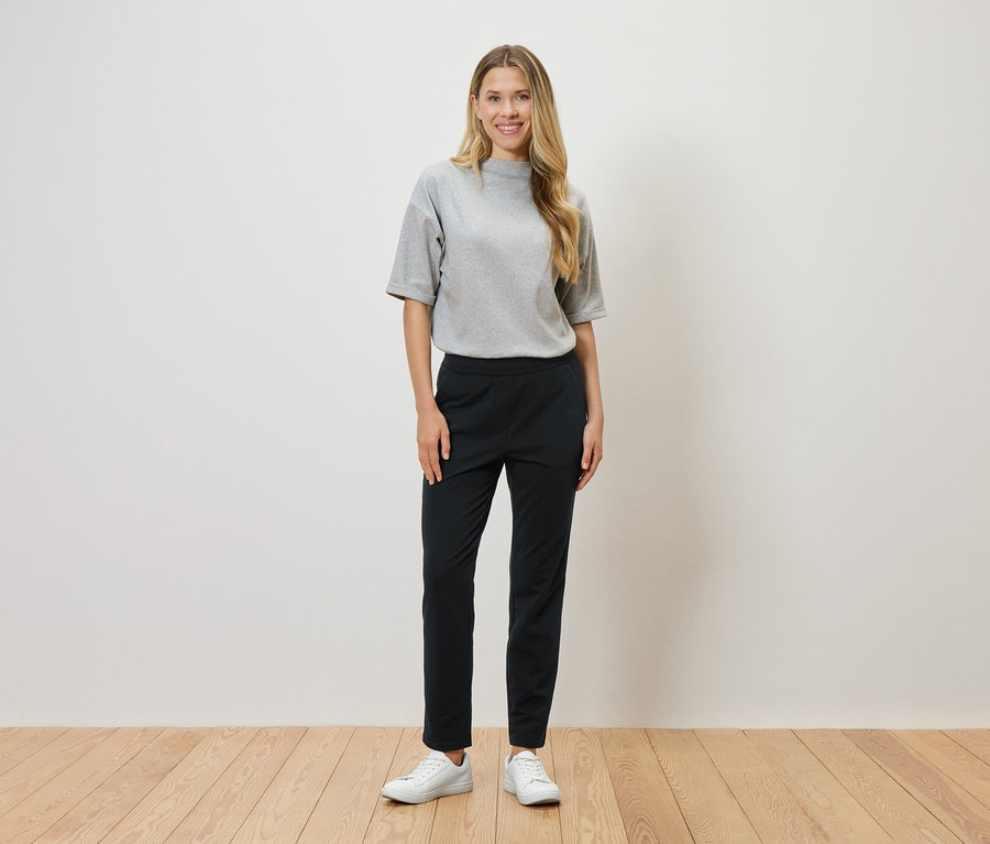 Kobieta o blond włosach stoi na tle białej ściany, ubrana w szarą koszulkę, czarne spodnie i białe tenisówki.