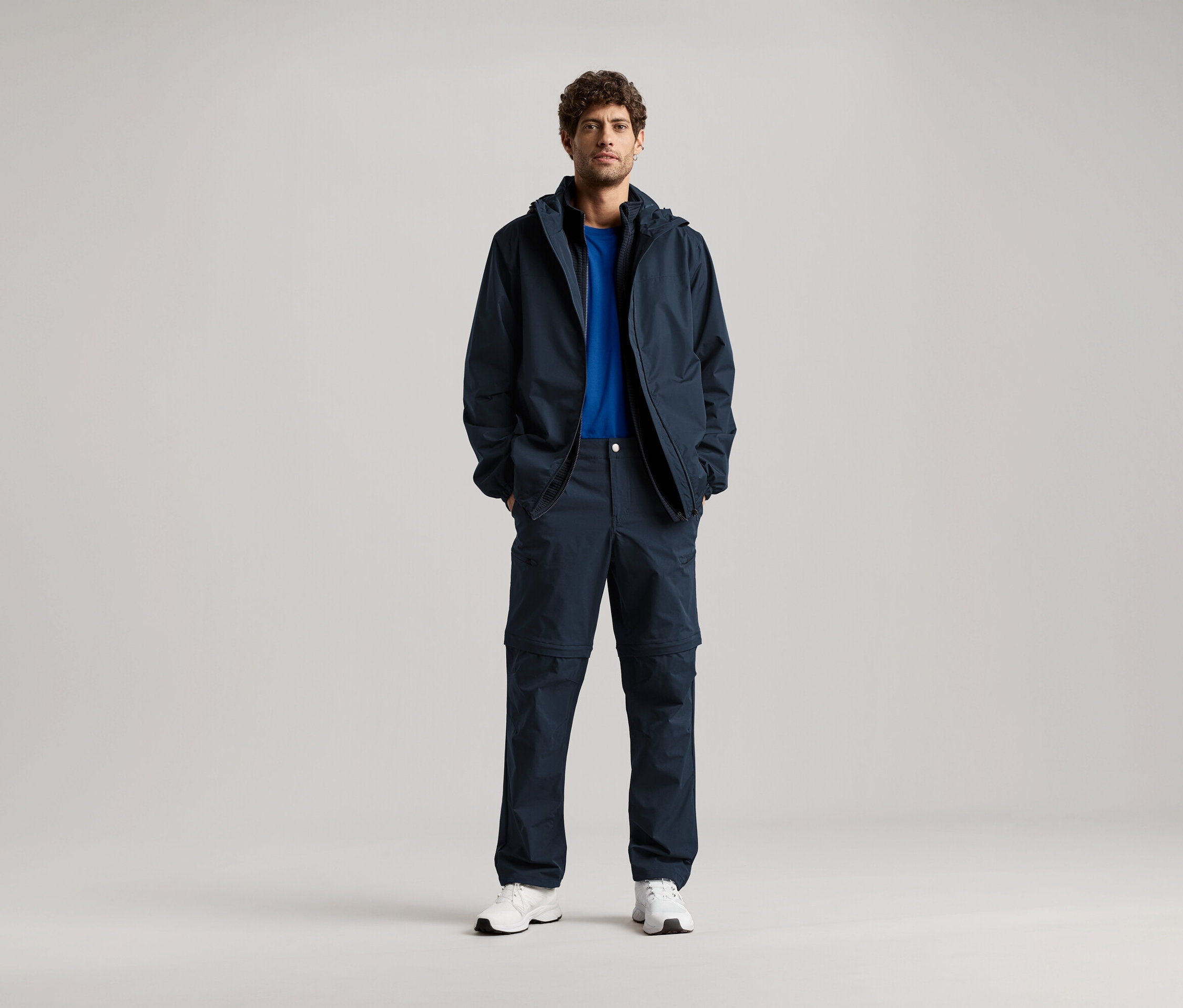 Mężczyzna stoi z rękami w kieszeniach granatowej kurtki i pasujących spodni, ma na sobie niebieski T-shirt i białe trampki.