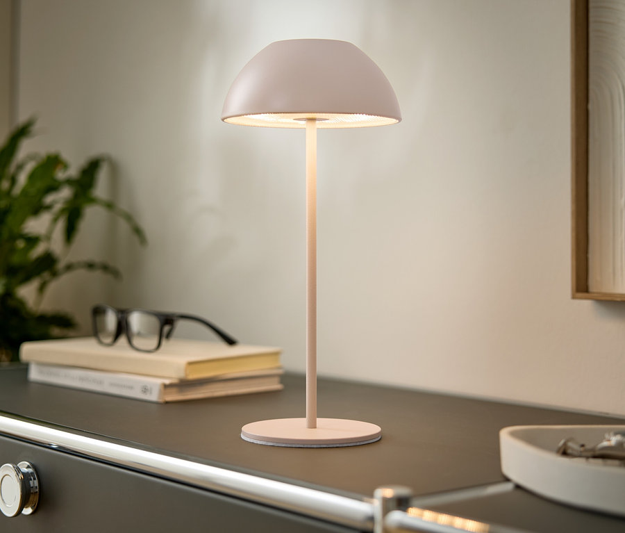 Na komodzie stoi różowa lampa, książki i okulary. W tle widać roślinę i oprawiony obraz.