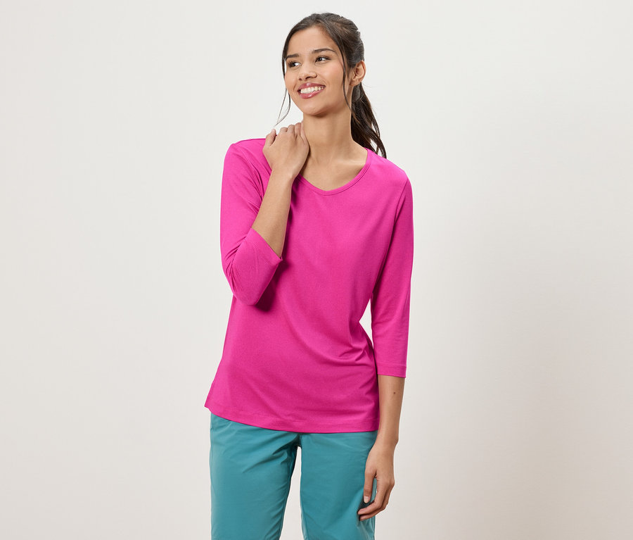 Kobieta z kucykiem ubrana w różową bluzkę z rękawami trzy czwarte i turkusowe spodnie.