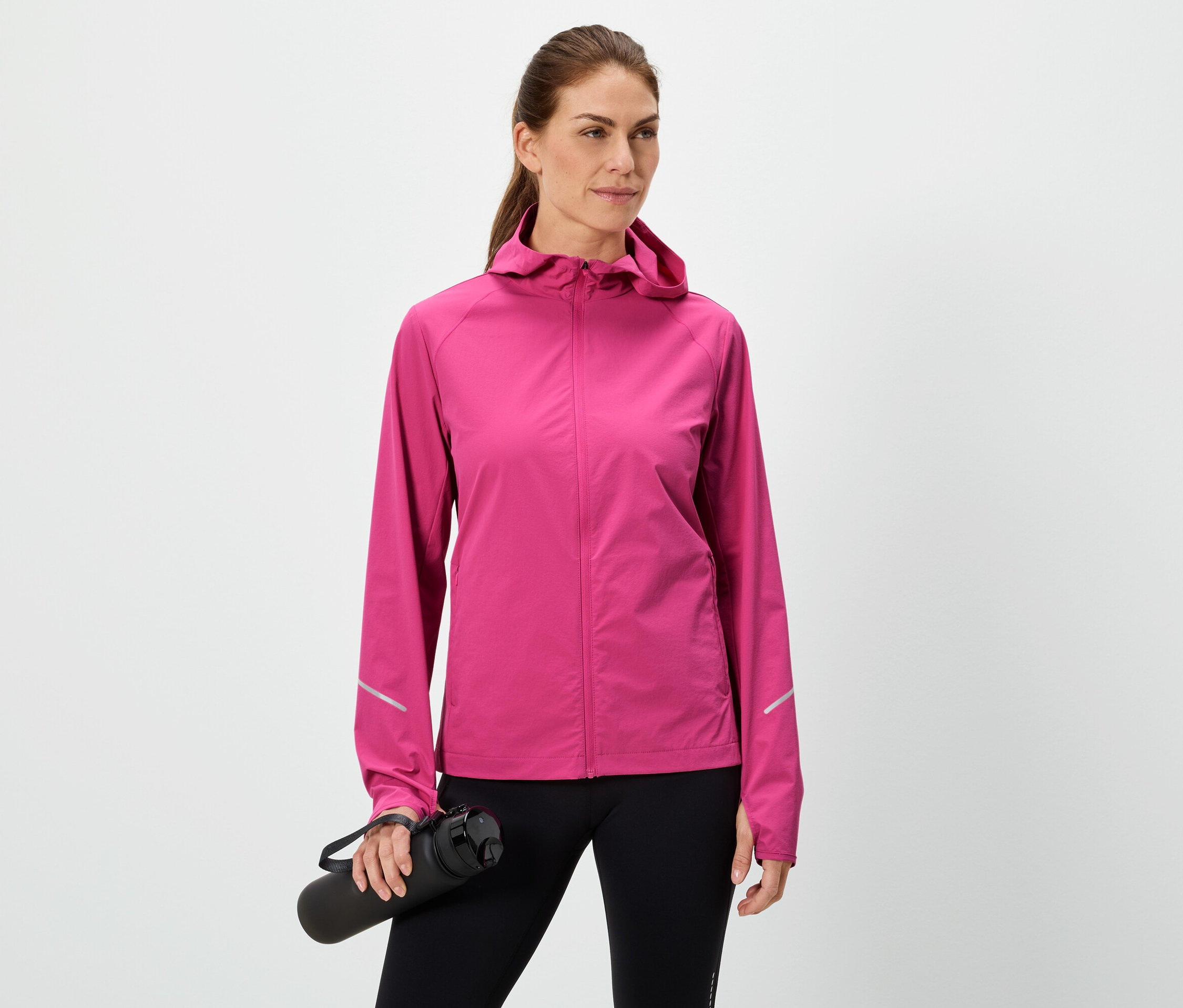 Kobieta trzyma czarną butelkę na wodę, ma na sobie różową kurtkę i czarne legginsy na białym tle.