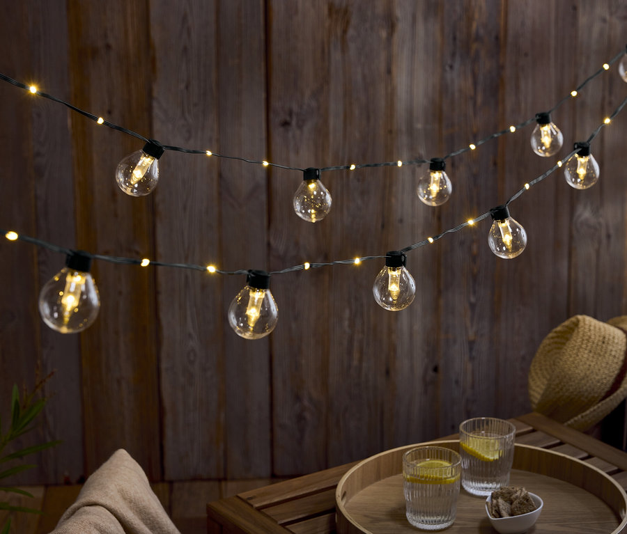 Dwa rzędy żarówek wiszą na drewnianej ścianie. Na drewnianym stole stoją dwie szklanki wody z cytryną i miska orzechów.