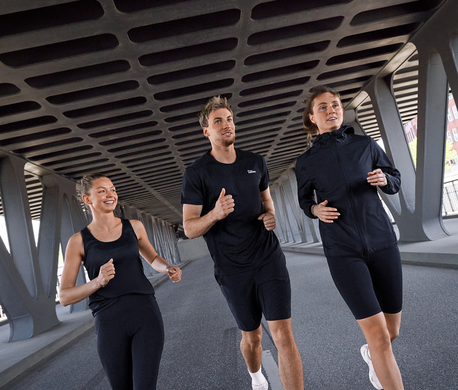 Trzy osoby, dwie kobiety i mężczyzna, biegną pod mostem. Kobieta po lewej ma na sobie czarny top sportowy i czarne legginsy sportowe „ActivePower”. Mężczyzna pośrodku ma czarną koszulkę sportową i czarne kolarki „ActivePower”. Kobieta po prawej ma czarną kurtkę wiatrówkę „packable” i czarne kolarki „ActivePower”.
