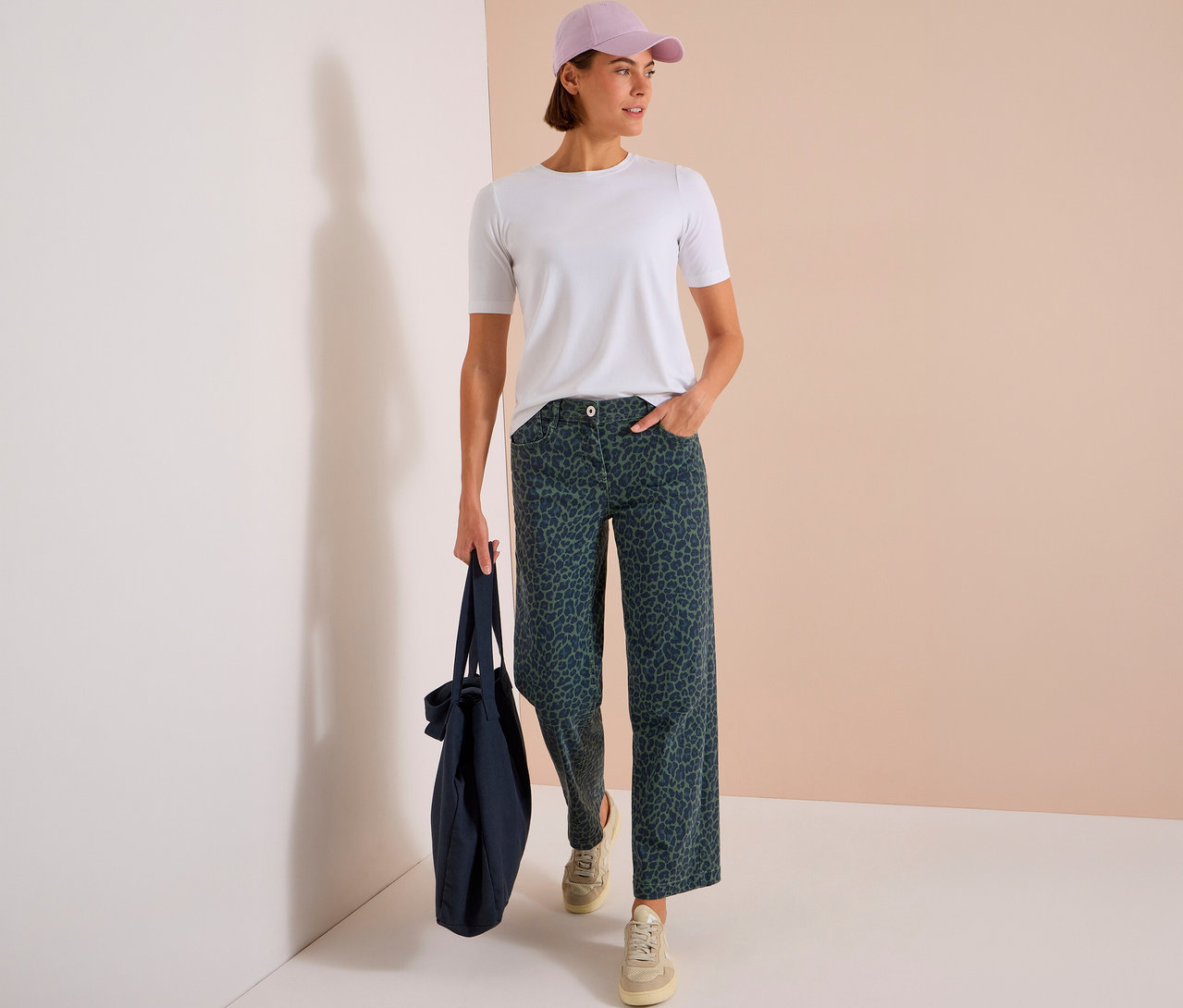 Stojąca kobieta w białym T-shircie, jasnofioletowej czapce i ciemnozielonych dżinsach CECIL w panterkę, trzymająca granatową torbę.
