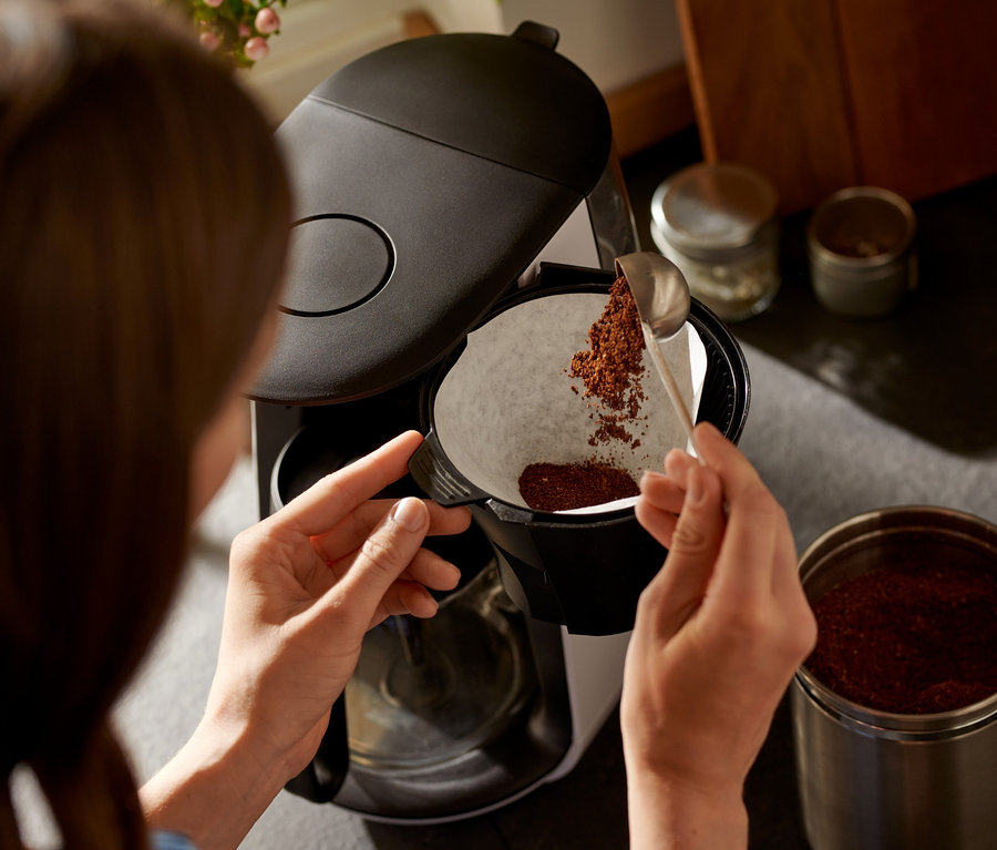 Kobieta wsypuje kawę łyżką do białego filtra ekspresu przelewowego »Let's Brew«.
