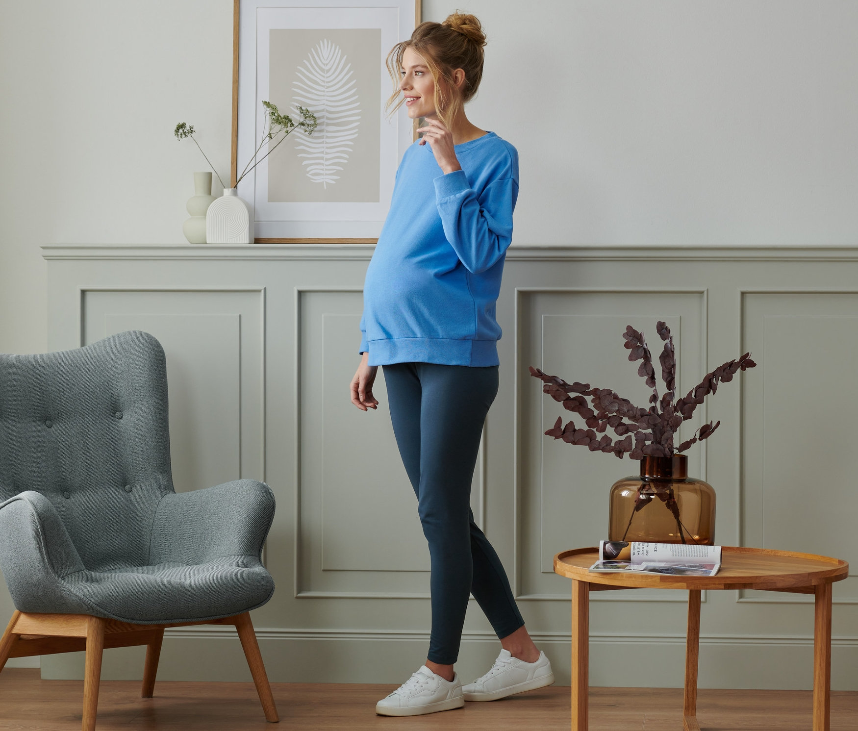Kobieta w ciąży stoi w pokoju, ubrana w niebieski top i legginsy ciążowe, 2 pary.