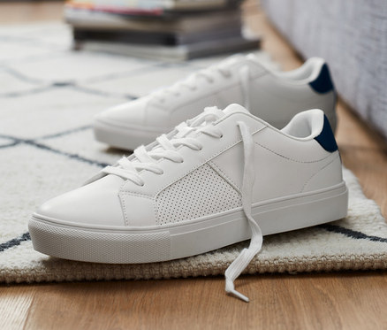 Dwa buty typu sneaker leżą na podłodze obok szarej kanapy.
