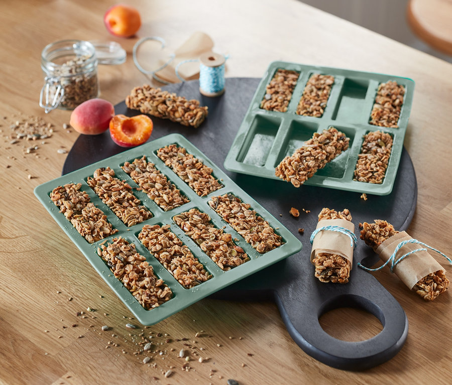 Dwie zielone formy do pieczenia batonów muesli, 2 sztuki, leżą obok siebie na stole. W jednej formie znajduje się pięć batonów, w drugiej osiem.