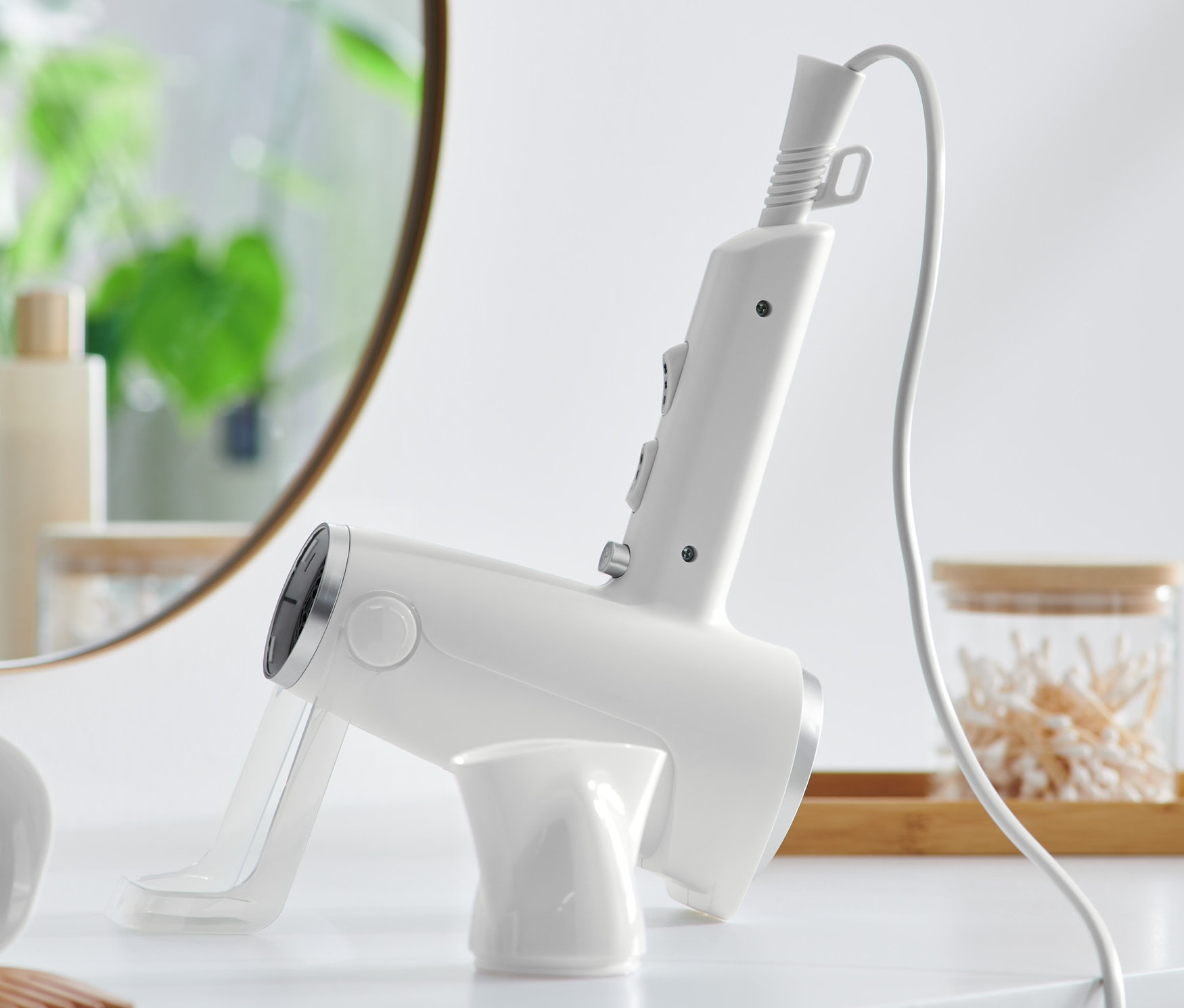 Biała suszarka do włosów ze stojakiem na białym stole. W tle lustro i kosmetyki.