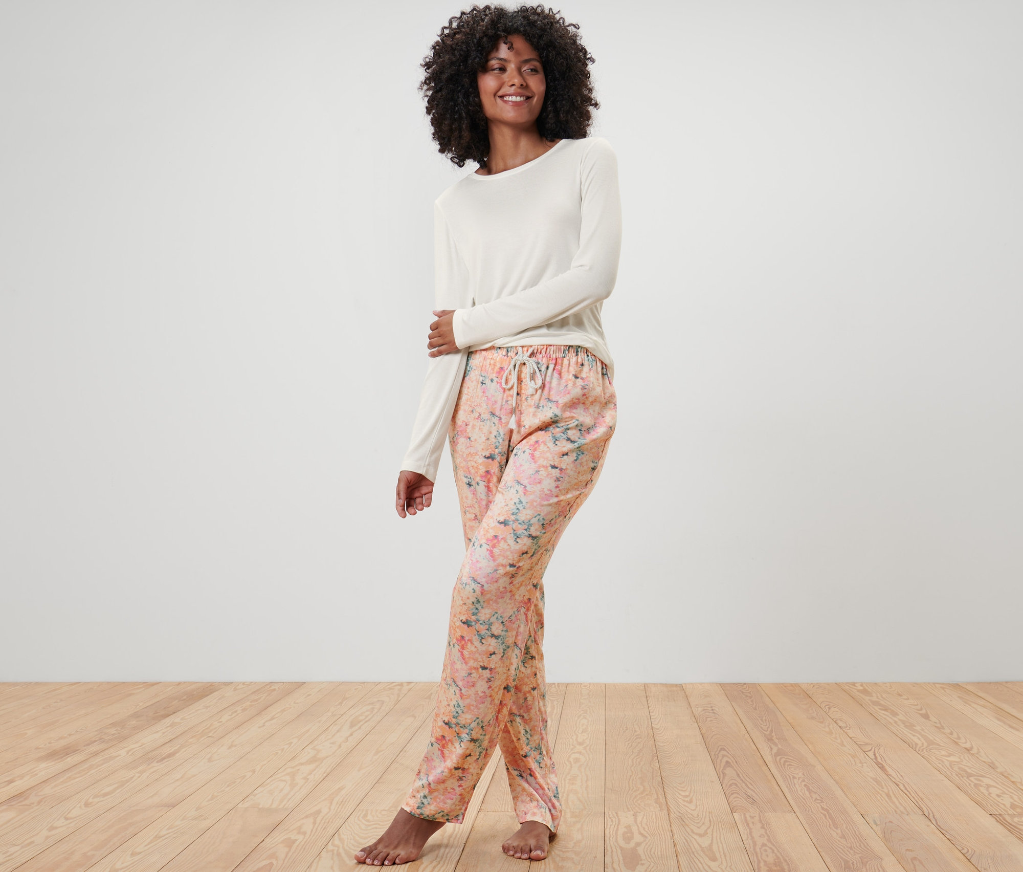 Kobieta w piżamie w kwiatowe spodnie i kremową górę stoi na drewnianej podłodze. Piżama.