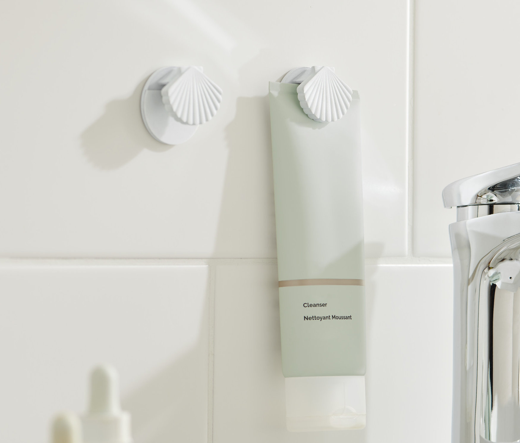 Dwa uchwyty na tubkę przymocowane do białych płytek, w jednym z nich znajduje się zielonkawa tubka.