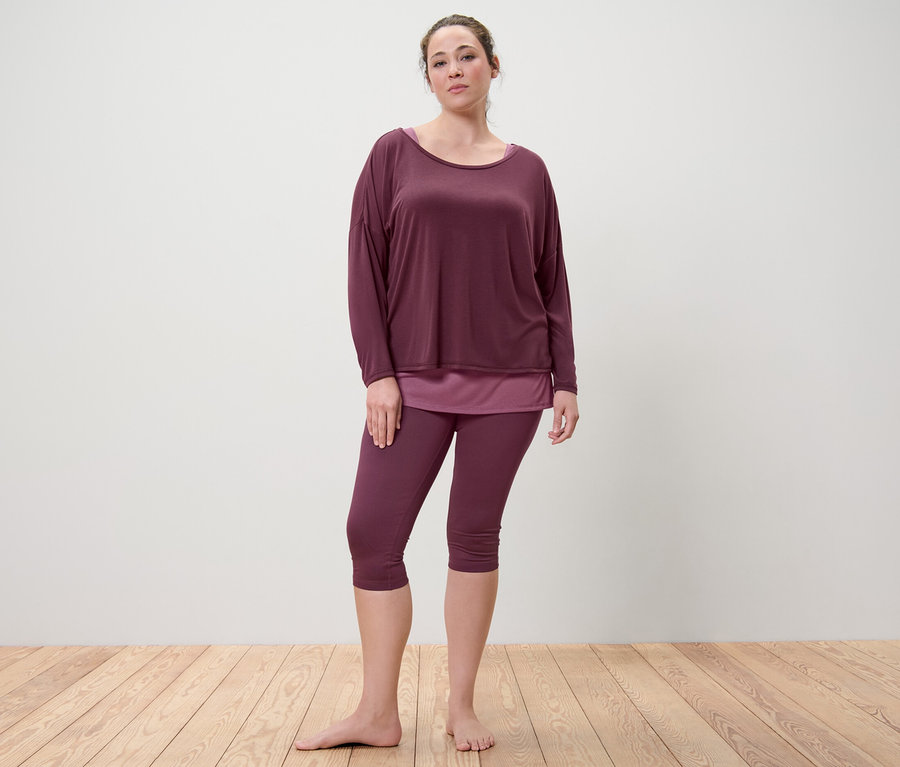 Kobieta stoi boso i ma na sobie śliwkową koszulkę sportową 2 w 1 i śliwkowe legginsy sportowe o długości 3/4.
