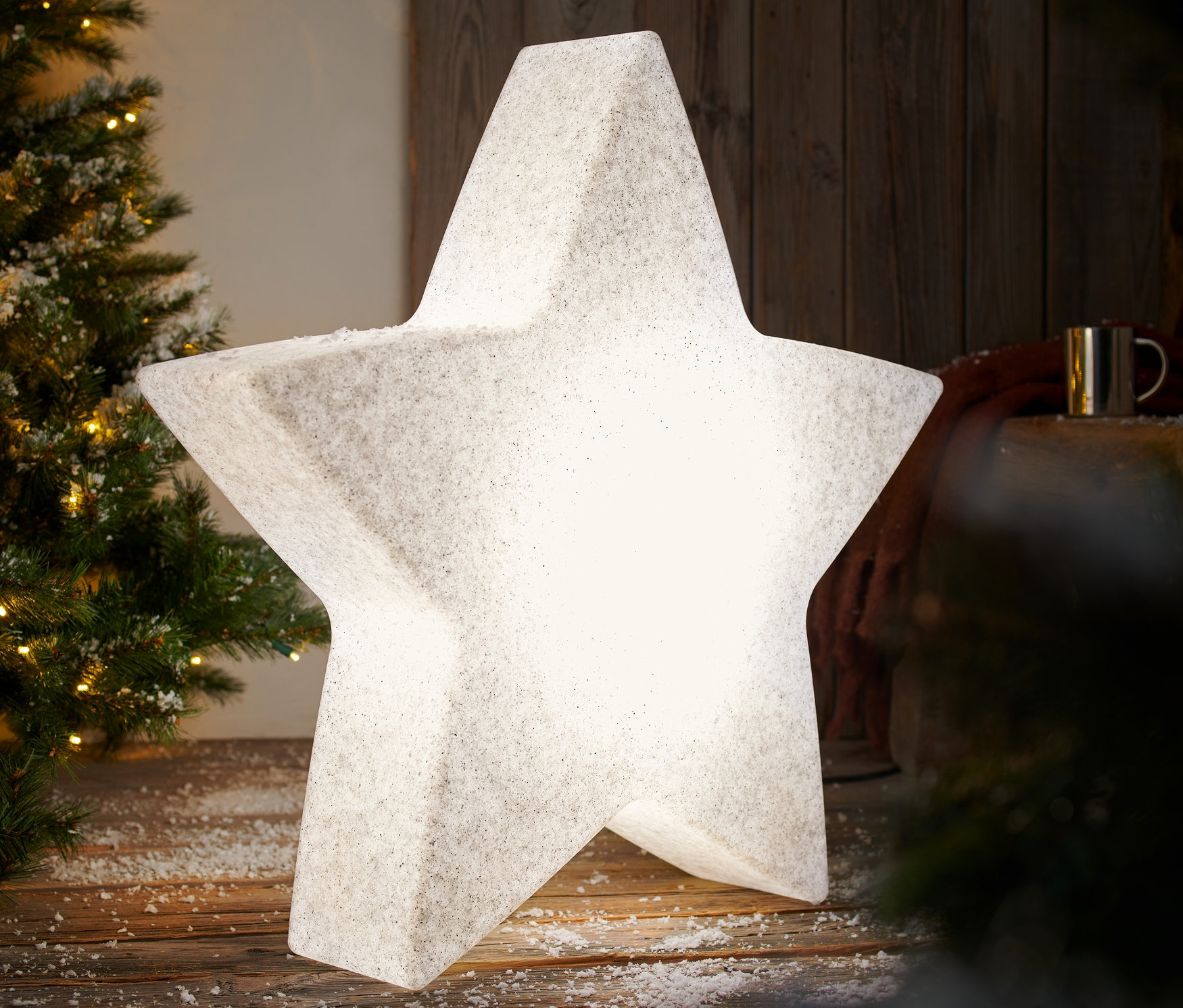 Świecąca inteligentna lampa ogrodowa »Gwiazda«, L, stoi na drewnianej podłodze pokrytej śniegiem obok choinki.