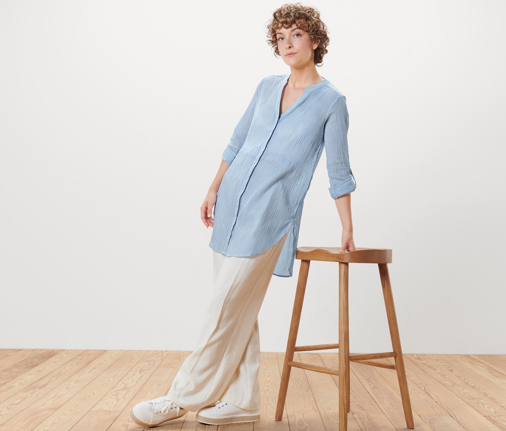 Kobieta oparta o drewniany stołek ma na sobie długą bluzkę w prążki.