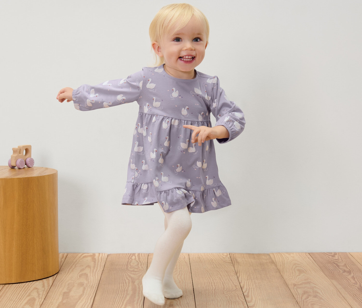 Dziewczynka w sukience w łabędzie i rajstopach stoi obok drewnianego stolika z zabawką.