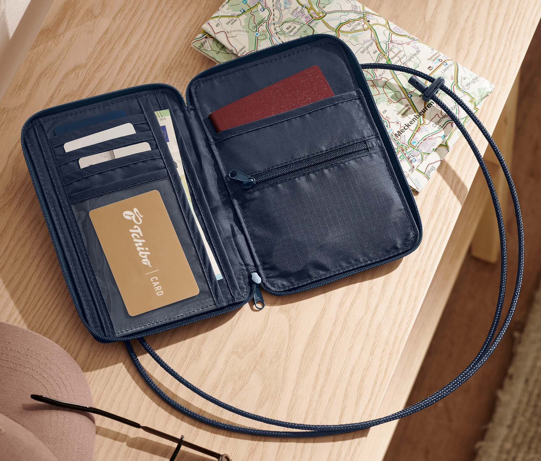 Otwarta, niebieska antykradzieżowa torebka na szyję leży na jasnej drewnianej powierzchni obok mapy, kluczy, kapelusza i okularów przeciwsłonecznych.