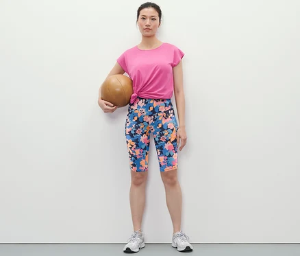 Kobieta trzyma piłkę lekarską i ma na sobie kolarki z nadrukiem na całej powierzchni, t-shirt i buty sportowe.