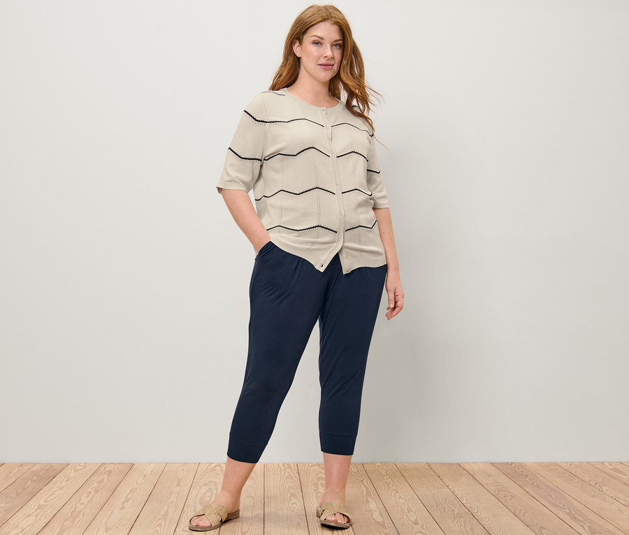 Kobieta pozuje w kardiganie z cienkiej dzianiny z ażurowym wzorem i spodniach dżersejowych o długości 3/4.