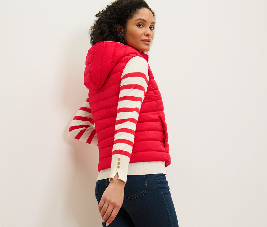 Kobieta patrzy przez ramię, ma na sobie kamizelkę pikowaną z kapturem, bluzkę w paski i dżinsy.