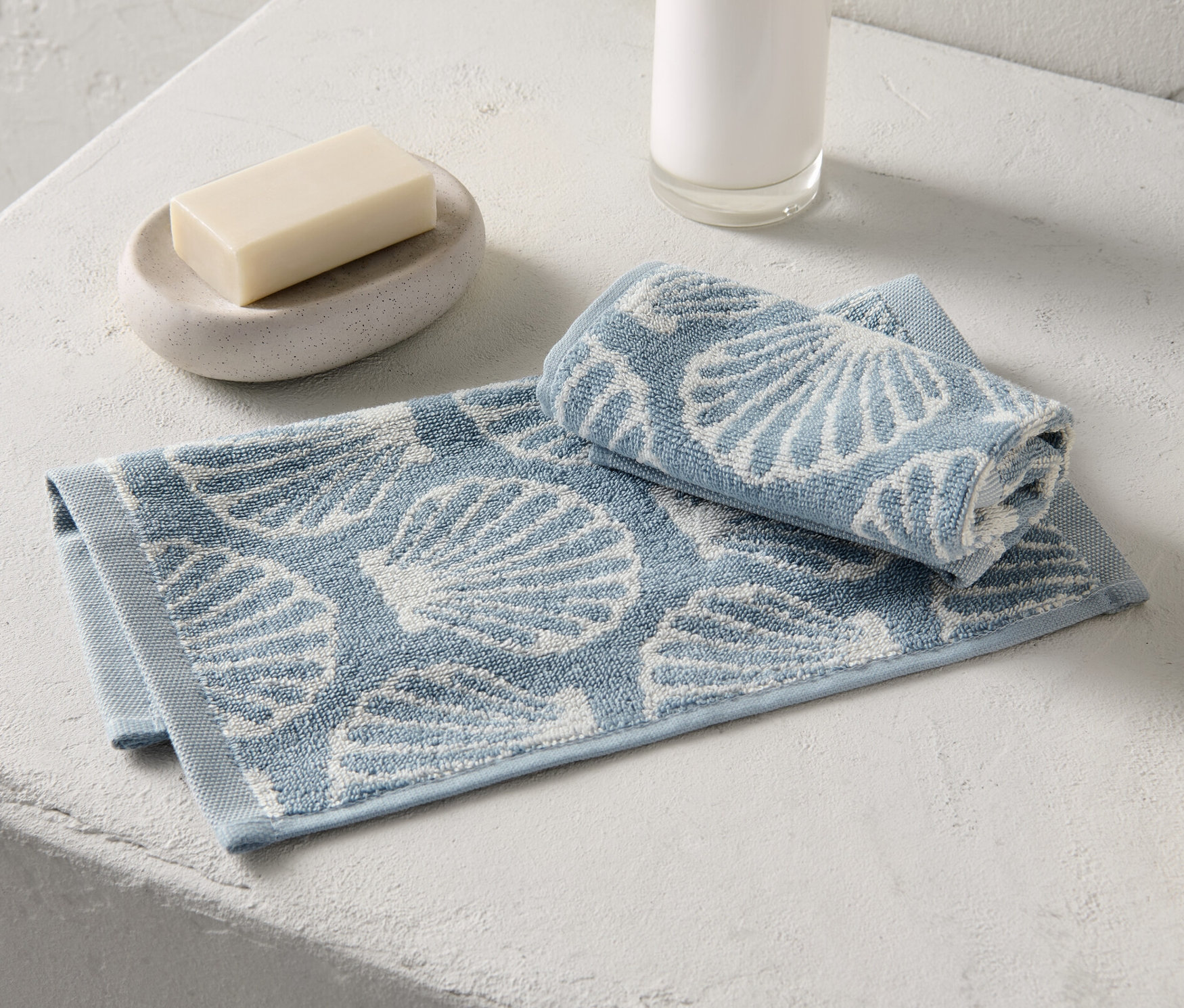 Dwa małe ręczniki ze wzorem muszelek leżą obok mydła i dozownika mydła.