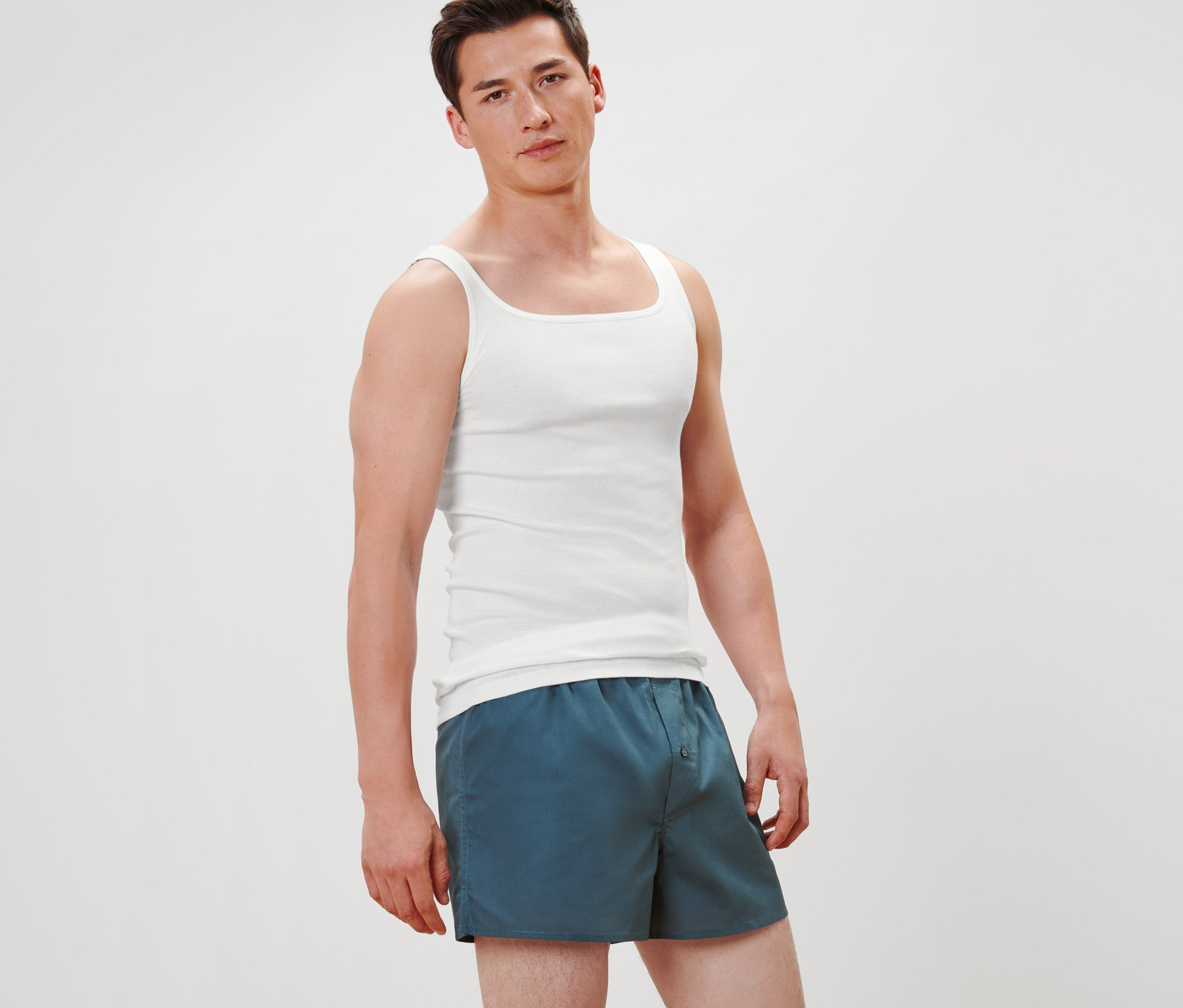 Mężczyzna stoi i ma na sobie 2 wysokiej jakości białe podkoszulki z dzianiny prążkowanej i niebieskie szorty.