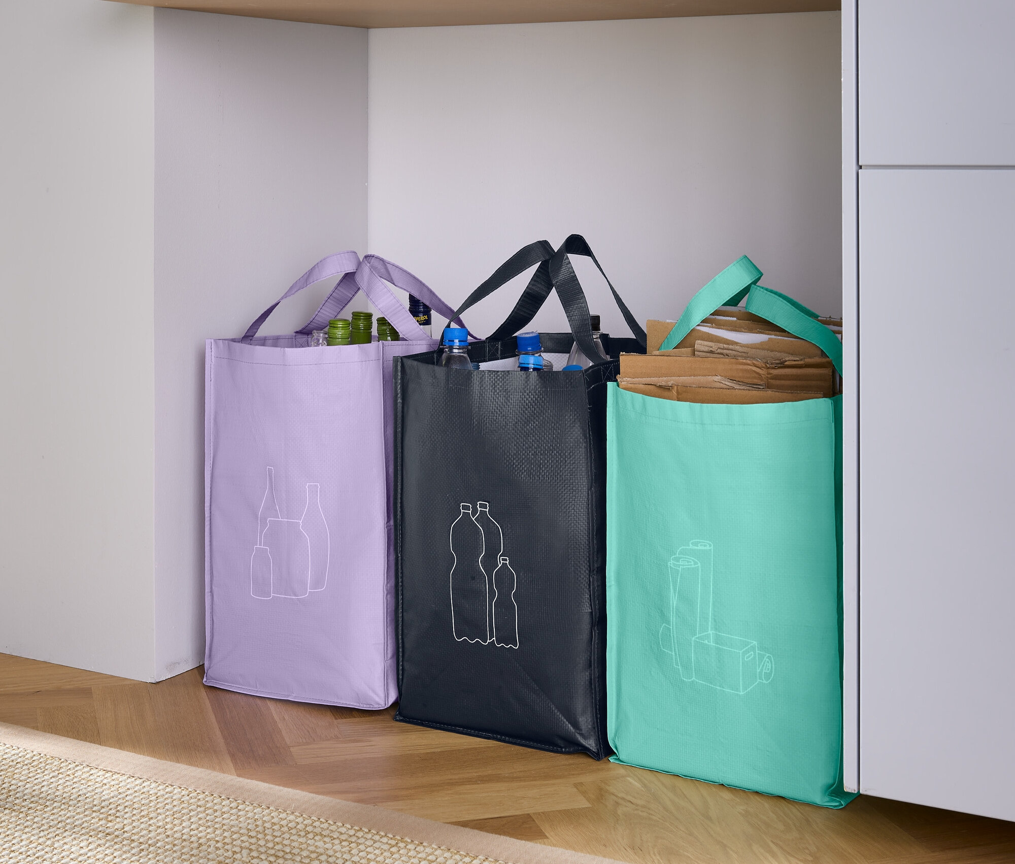 Trzy torby na zakupy stoją obok siebie: fioletowa torba z rysunkiem owoców, czarna torba z rysunkiem butelek i turkusowa torba z rysunkiem szklanki.