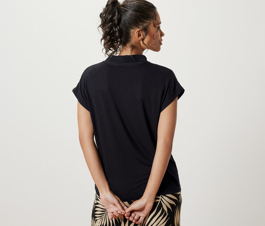 Kobieta tyłem do kamery ma na sobie czarną bluzkę z dekoltem w serek i spodnie we wzór liści.