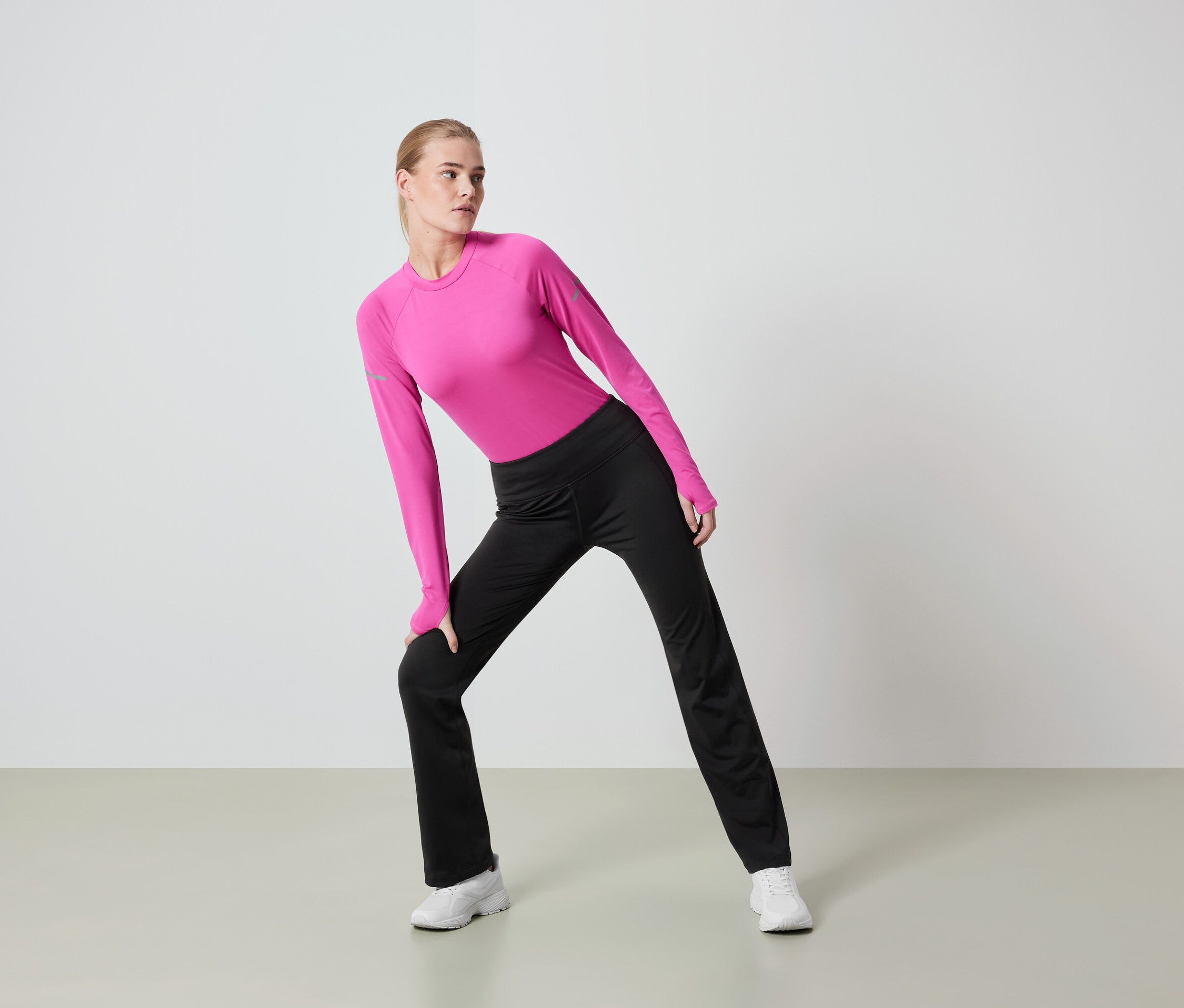 Kobieta o blond włosach rozciąga się w różowym topie z długim rękawem i czarnych spodniach.
