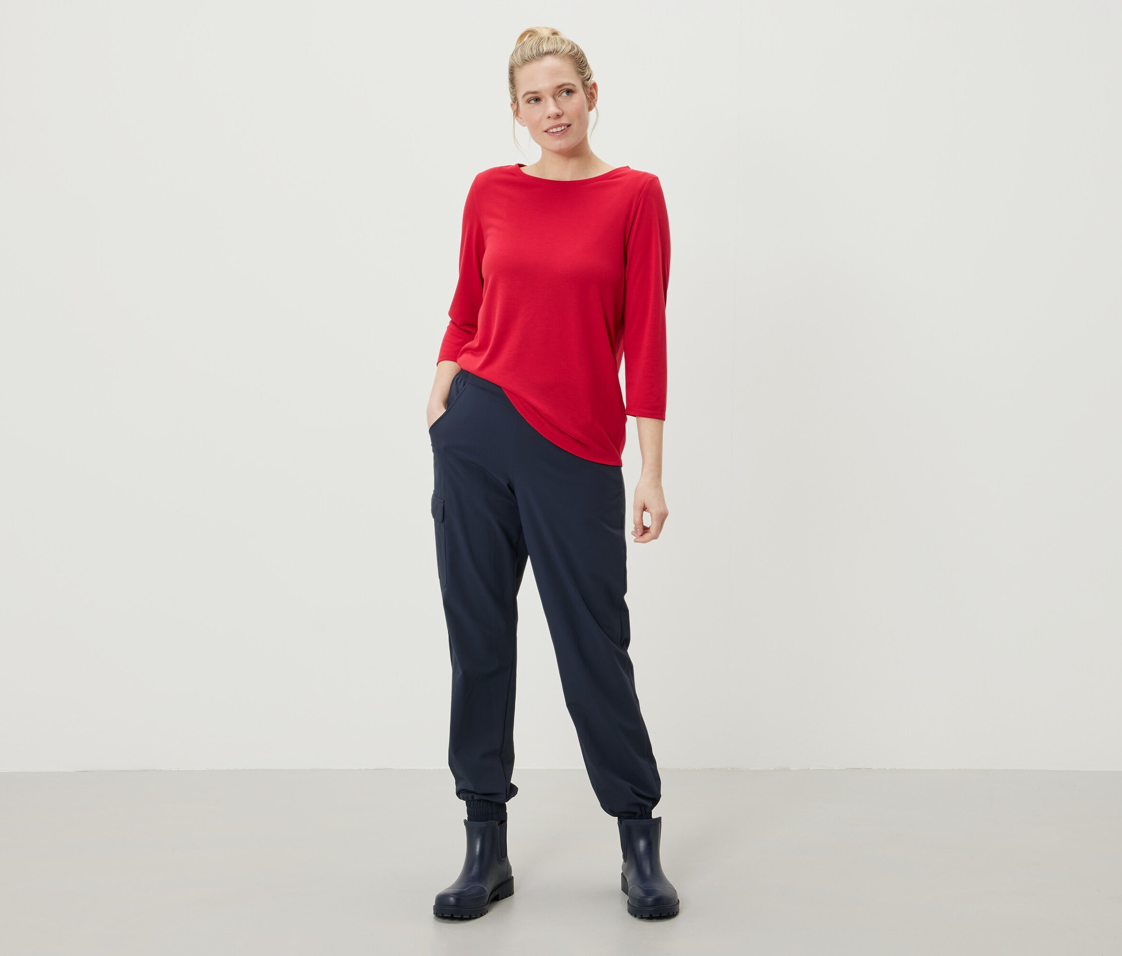 Kobieta stoi z rękami w kieszeniach granatowych spodni, ma na sobie czerwoną bluzkę z rękawami trzy czwarte i granatowe kalosze.