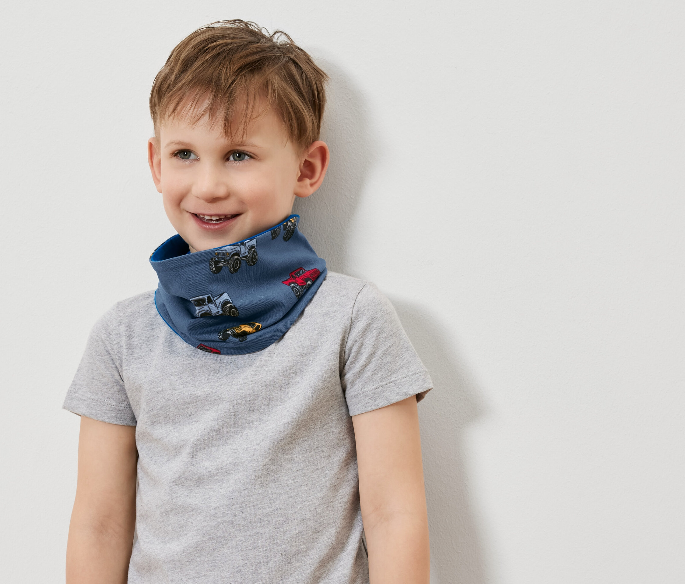 Uśmiechnięty chłopiec w niebieskim kominie z nadrukiem samochodów i szarej koszulce.